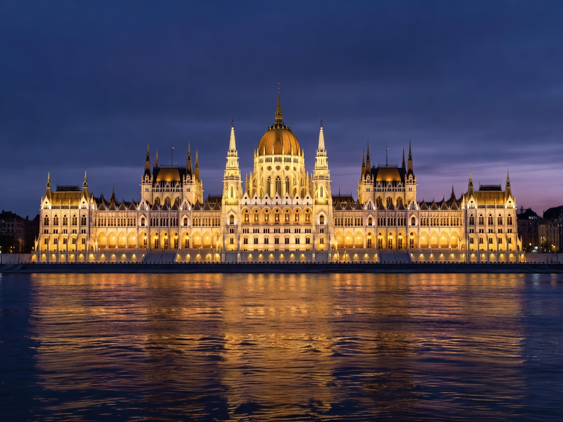 Full view of glowing Hungarian Parliament building from river water at dusk
